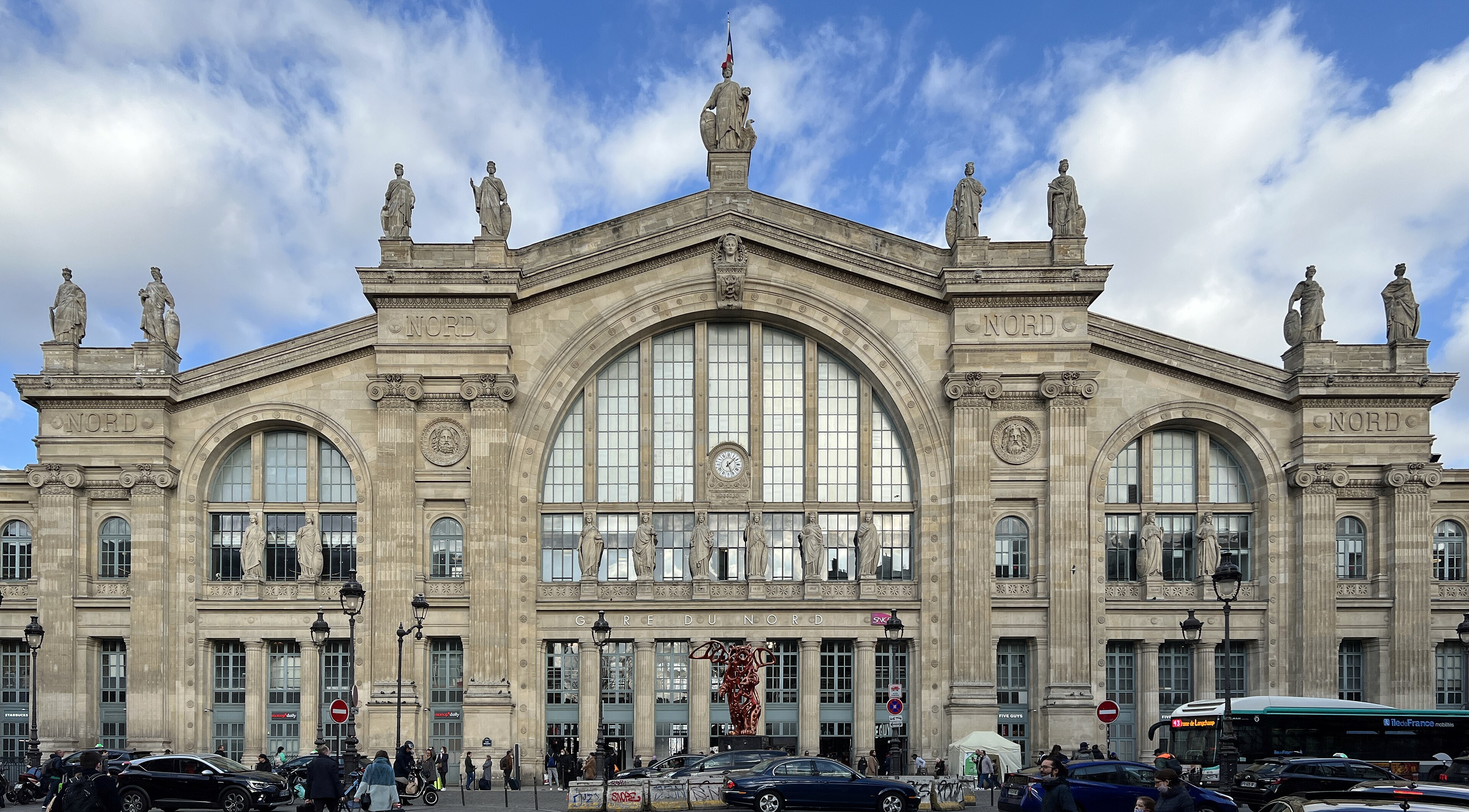 Façade de la gare du Nord, statues, grande verrière, sculpture rouge à l’entrée, bus et voitures, janvier 2022.