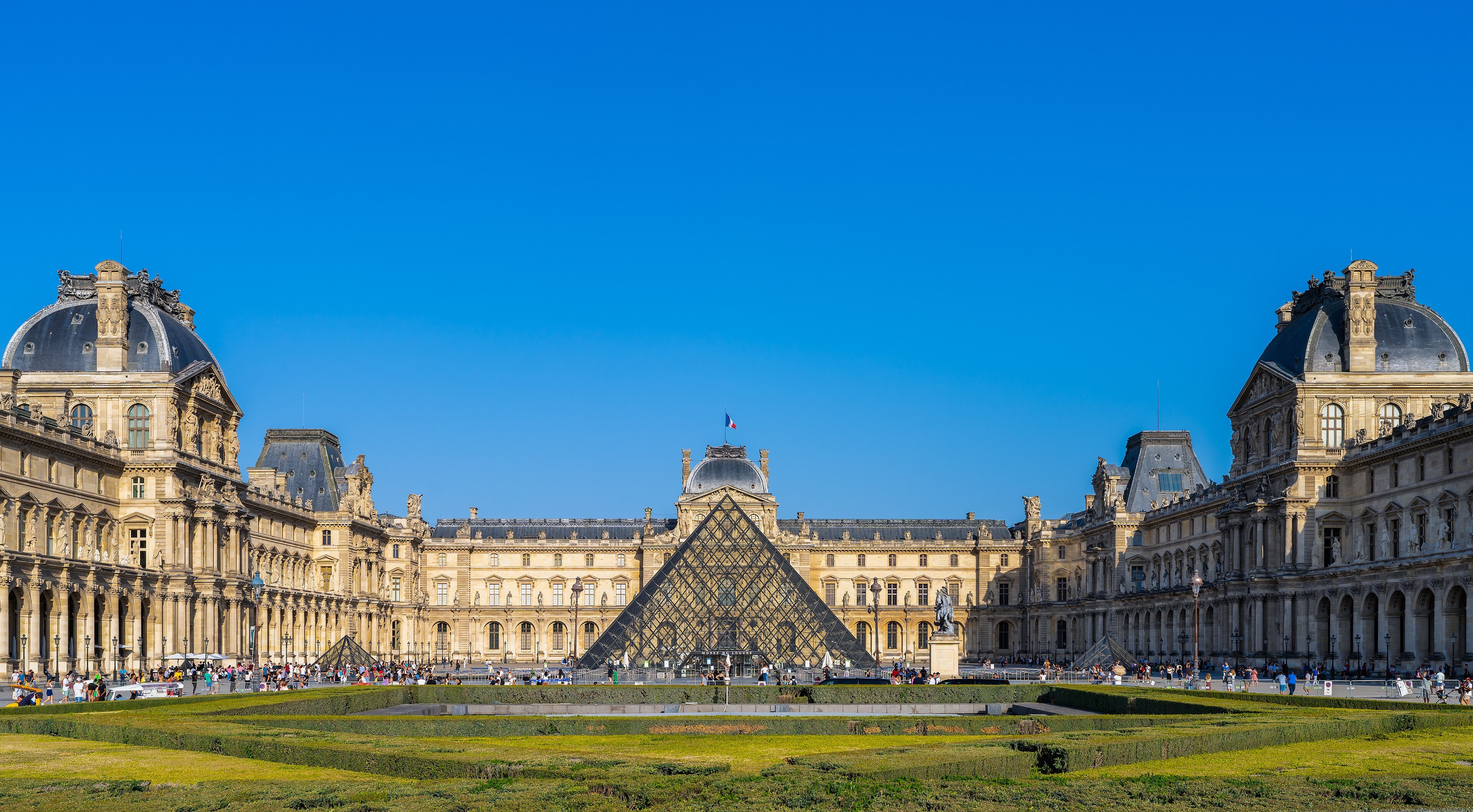 Cour Napoléon du Louvre : pyramide de verre, parterres, ailes et foule sous un ciel bleu.
